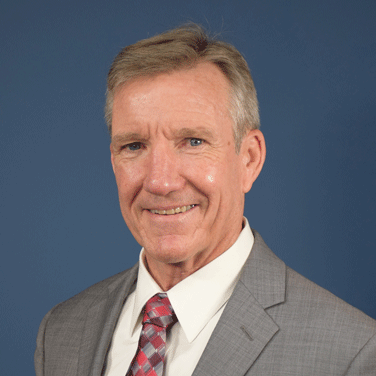 Image of Hawk Carlisle; Carlisle is seated in front of a blue background. He wears a gray suit, multi-colored tie, has graying hair, and is smiling showing his teeth.