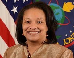 Image of Janice Glover-Jones, an African American woman with brown hair cropped around her shoulders. She is seated in front of the U.S. flag, has on a gold blazer and has an open mouth smile.