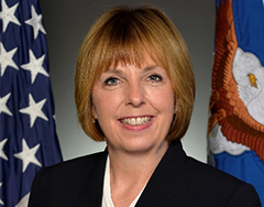 Image of Terri Sanchez; Sanchez has light blond hair and is seated before an American and U.S. Air Force Flag. She has an open mouth smile, showing her teeth.