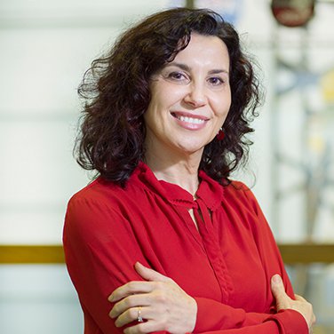 Image of Dr. Begoña Vila; Vila is standing with her arms crossed. She is smiling with her teeth showing, wears a red shirt, and has wavy dark hair that reaches her shoulders. 