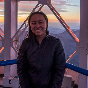 Dr. Christine Chen is standing on top of a high building overlooking a mountainous background. She is wearing a dark jacket, has dark hair, and is smiling with her teeth showing. 