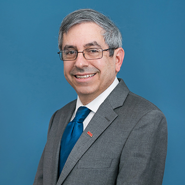 Dr. Mark Lewis stands in front of a blue background. He has a gray suit with a blue tie and an "NDIA" badge. He has gray-brown hair, glasses, and is smiling with his teeth showing. 
