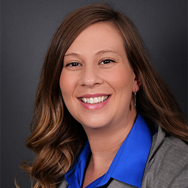 Erin Wolf is seated in front of a gray background, has a blue shirt and gray blazer on, has long curly brown hair and is smiling with her teeth showing.