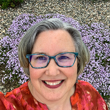 Jacqueline Maldonado is outside with purple and yellow flowers behind her. She has gray hair, a bright red shirt on, blue glasses, and is smiling with her mouth open.