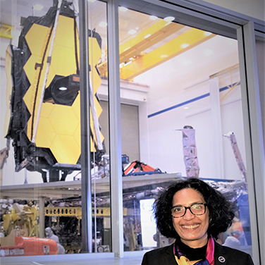 Bhavya Lal is standing before the James Webb Telescope. She has a dark shirt, dark glasses, and dark hair, and is smiling with her teeth showing. 