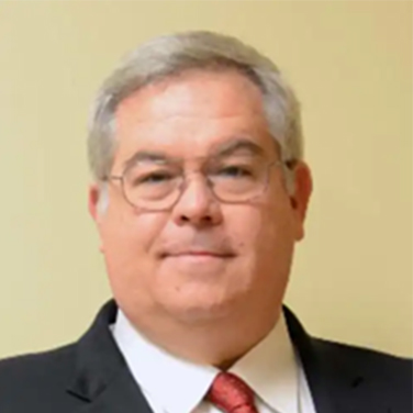 Image of Jim Moore. More is standing in front of a beige wall, has graying hair, round glasses, and has a closed mouth smile. He wears a dark suit with a red tie. 